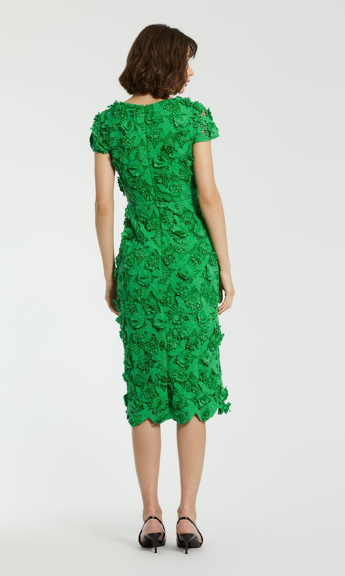 A woman stands facing away in a knee-length green Midi Lace Cocktail Dress by Mac Duggal 81022 and black heels, set against a plain white background.